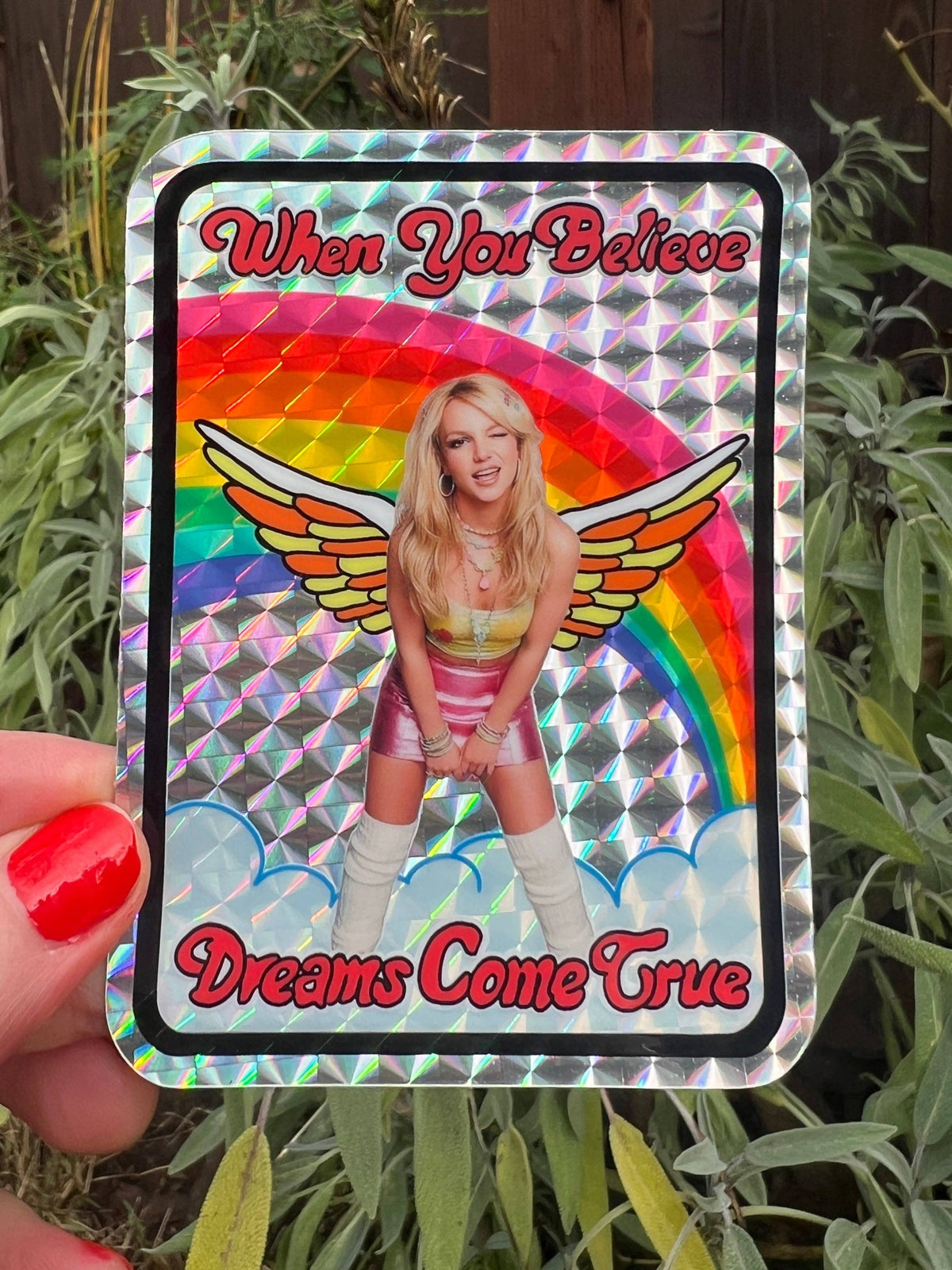 Rectangular holographic sticker with black frame, wings, rainbow arch, and “When You Believe / Dreams Come True”.
