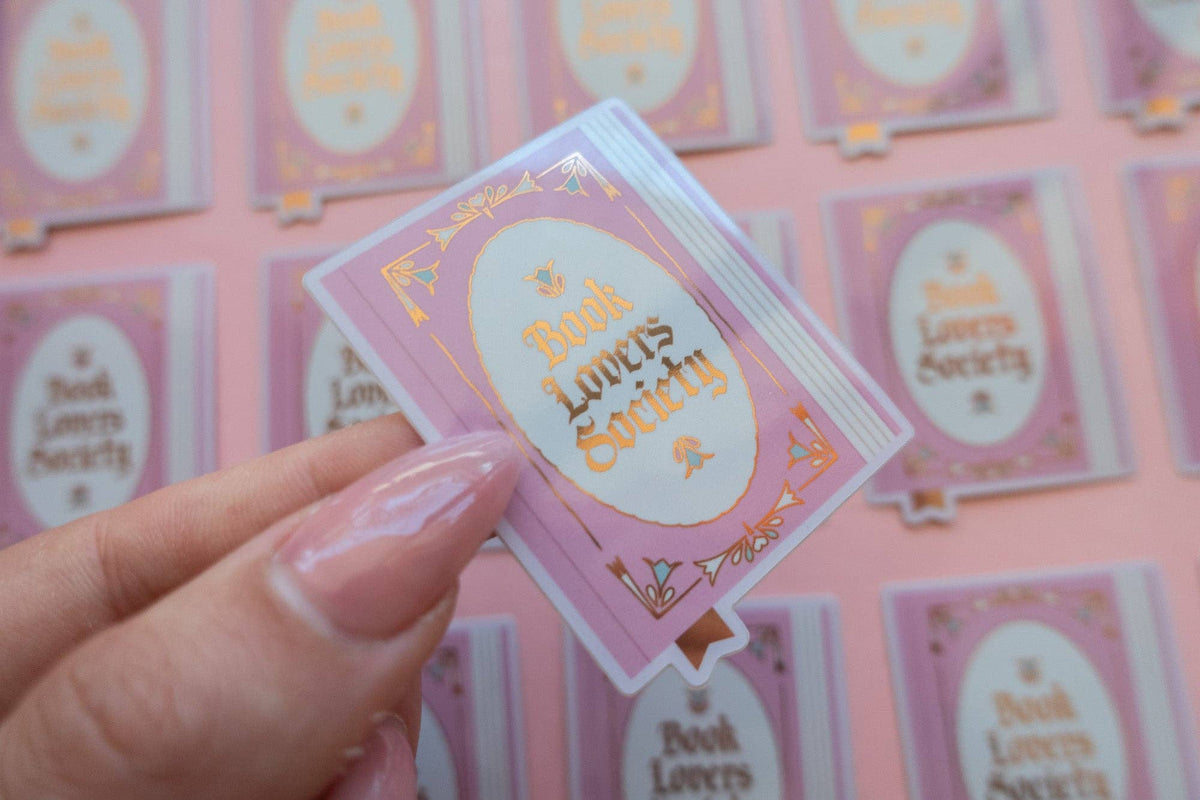 Hand holding vinyl sticker with light blue frame and gold "Book Lover's Society" lettering