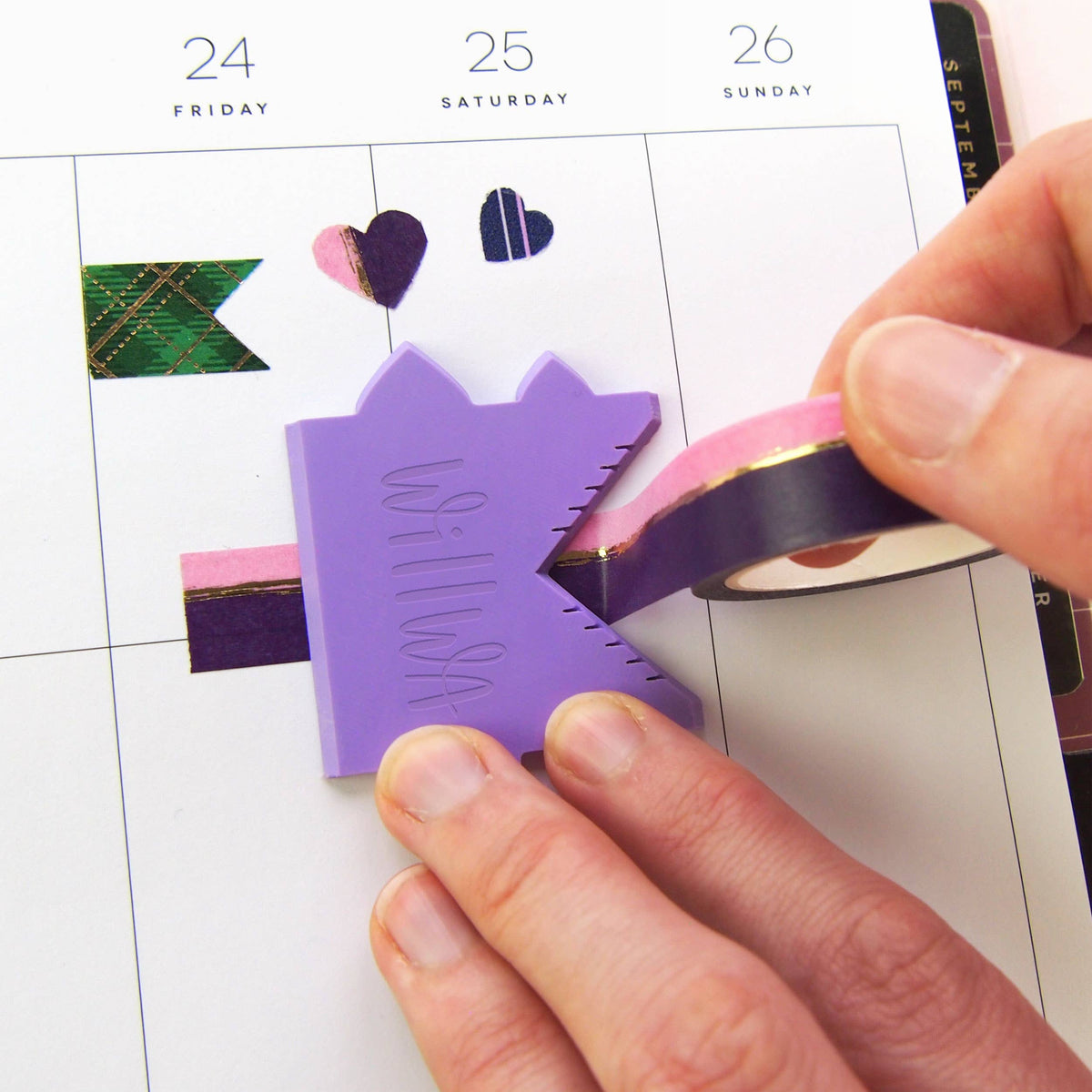 Hand pressing a purple house-shaped washi cutter onto a planner with dark tape strip