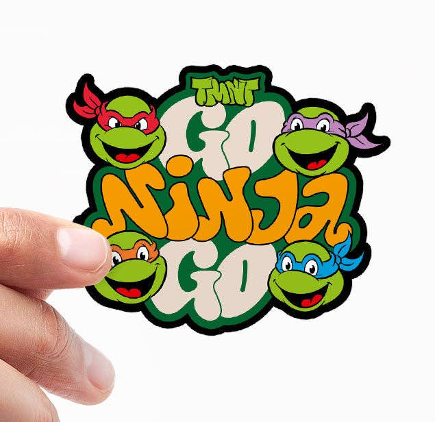 Cartoon ninja turtle vinyl sticker held by fingers, green faces with orange letters