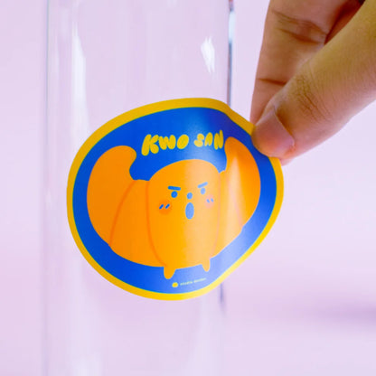 Vinyl croissant sticker held by hand, circular blue outline with orange character face