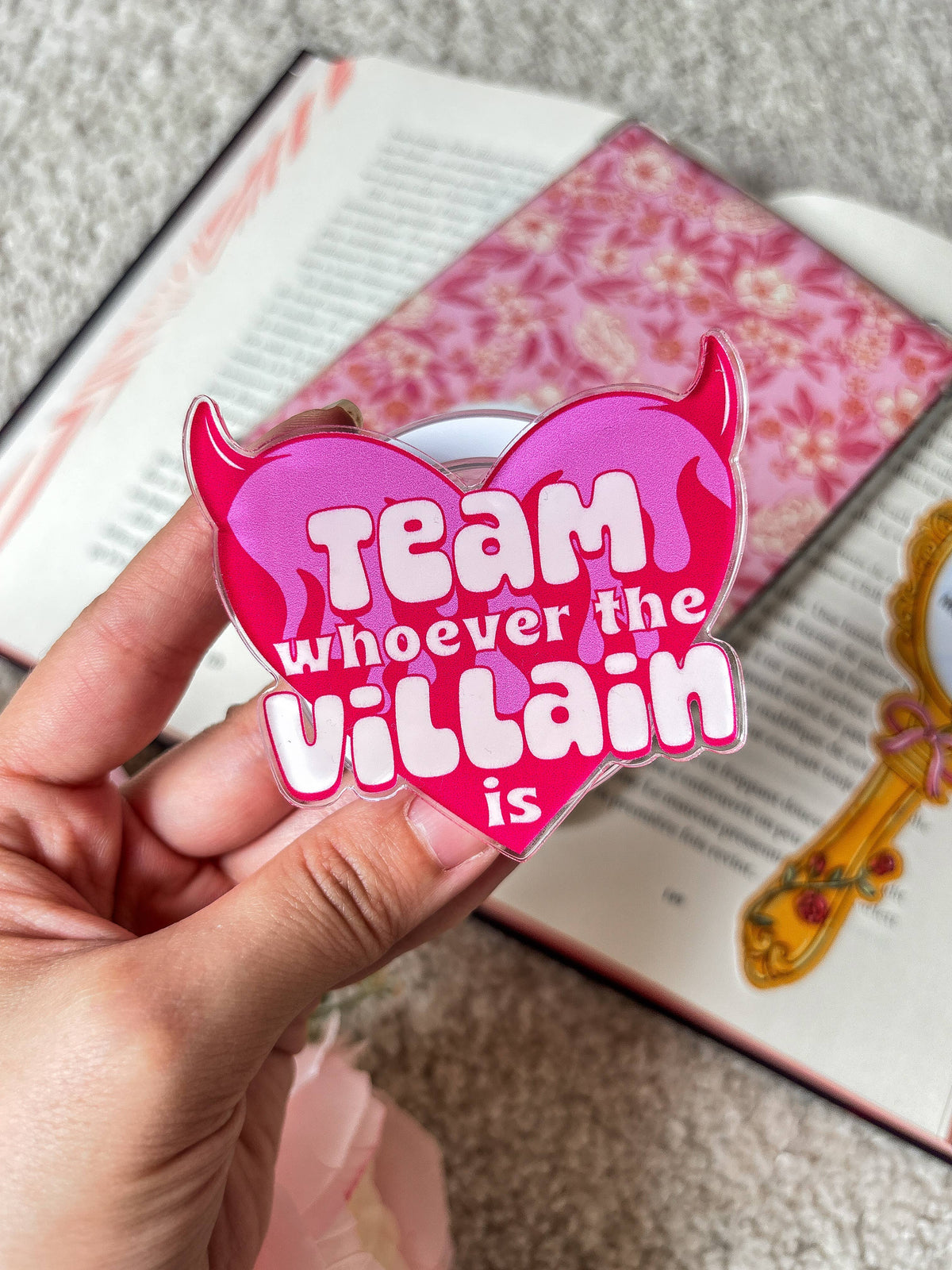 Hand holding pink devil heart magnetic grip over book, 'Team whoever the villain is'
