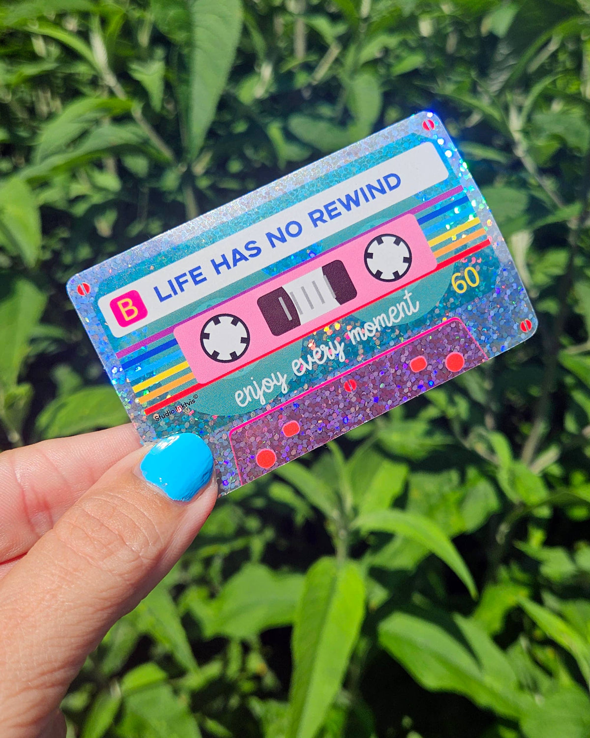 Glitter cassette tape sticker held in hand, blue and rainbow stripes; 'Life has no rewind'.