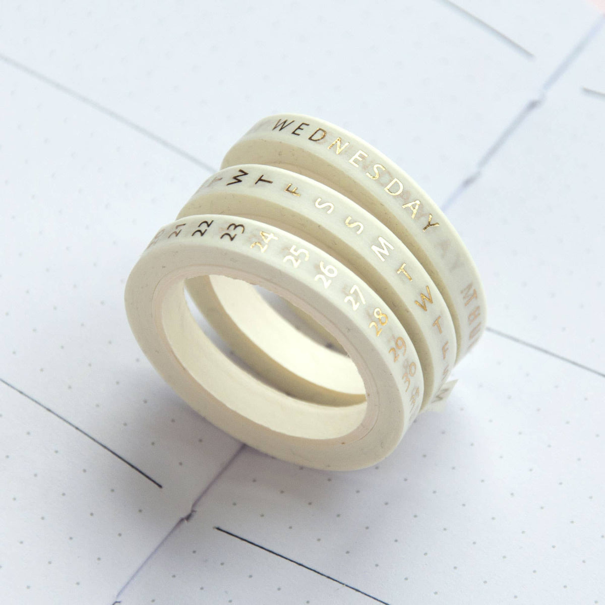 Stacked thin washi tape rolls with day names and date numbers in beige tones