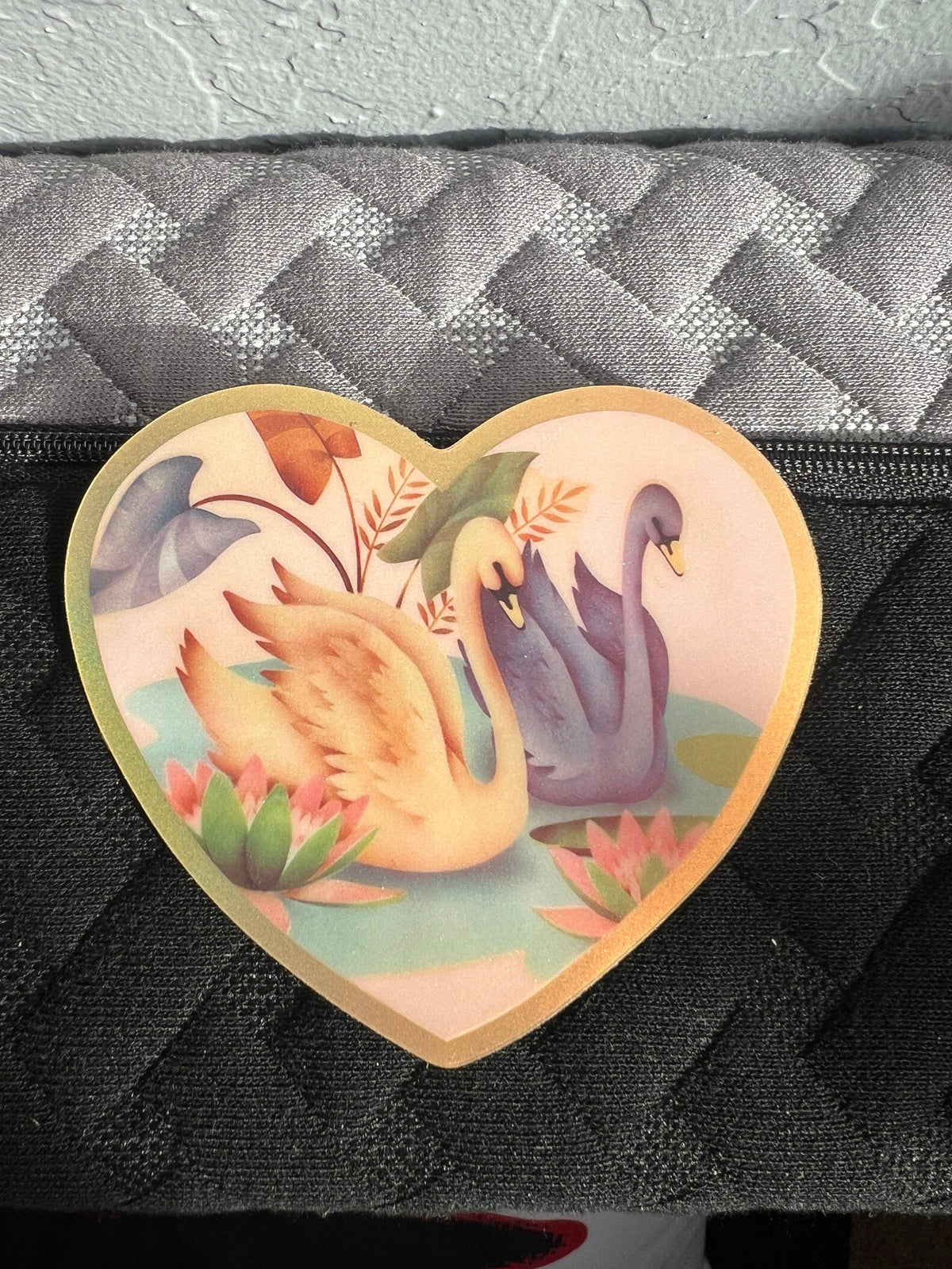 Heart-shaped Swan Friends sticker, 3 inch, showing swans and plants on a black bag