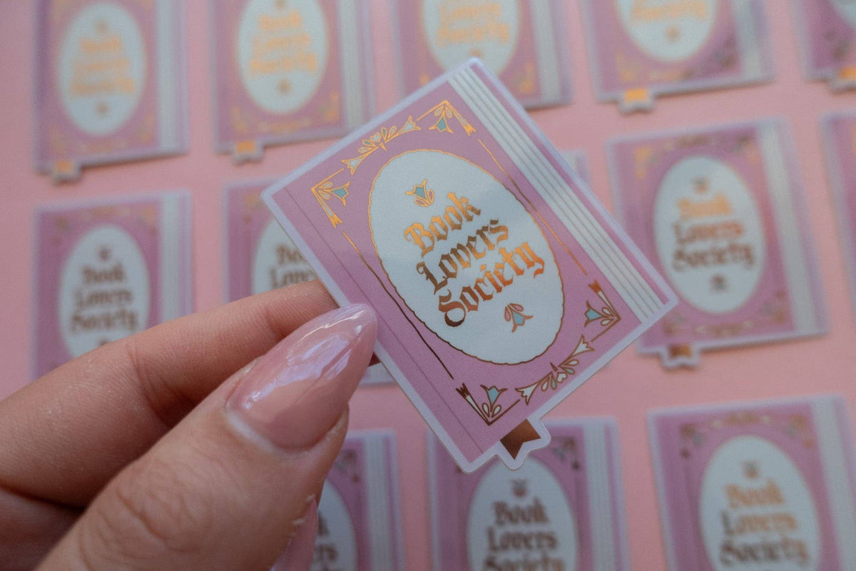 Hand holding a vinyl sticker with purple border and oval “Book Lovers Society” artwork