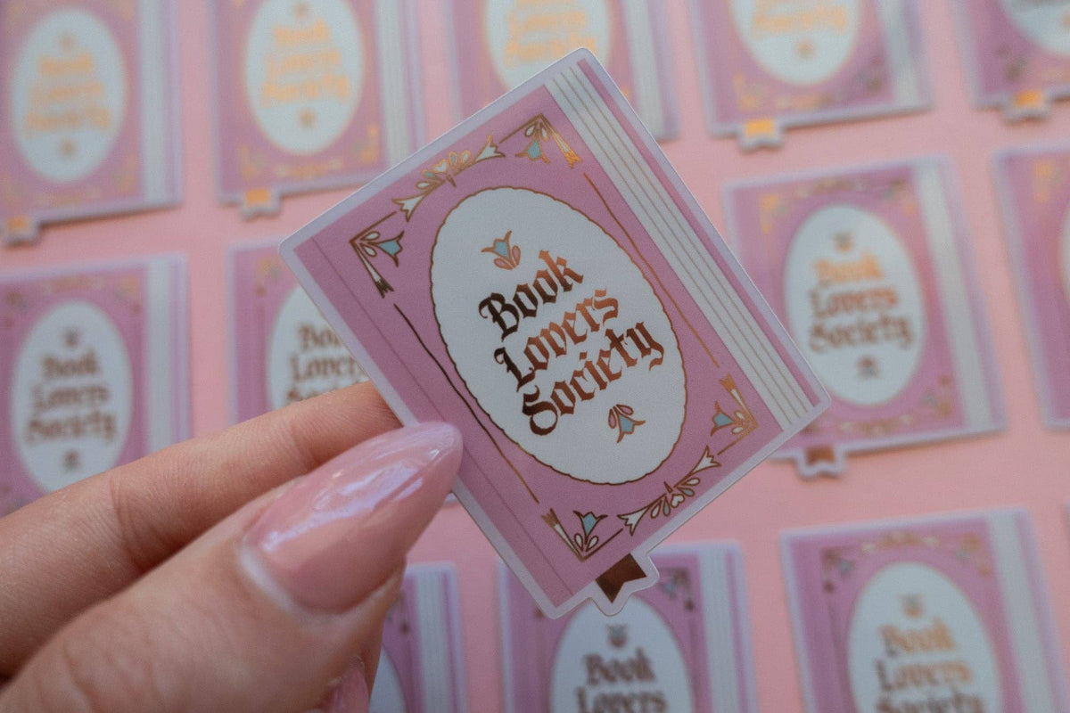 Hand holding a vinyl sticker with purple border and oval “Book Lovers Society” in gold