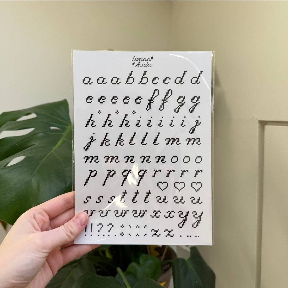 A5 sticker sheet held in hand with pixel letters, numbers, and heart icons in black