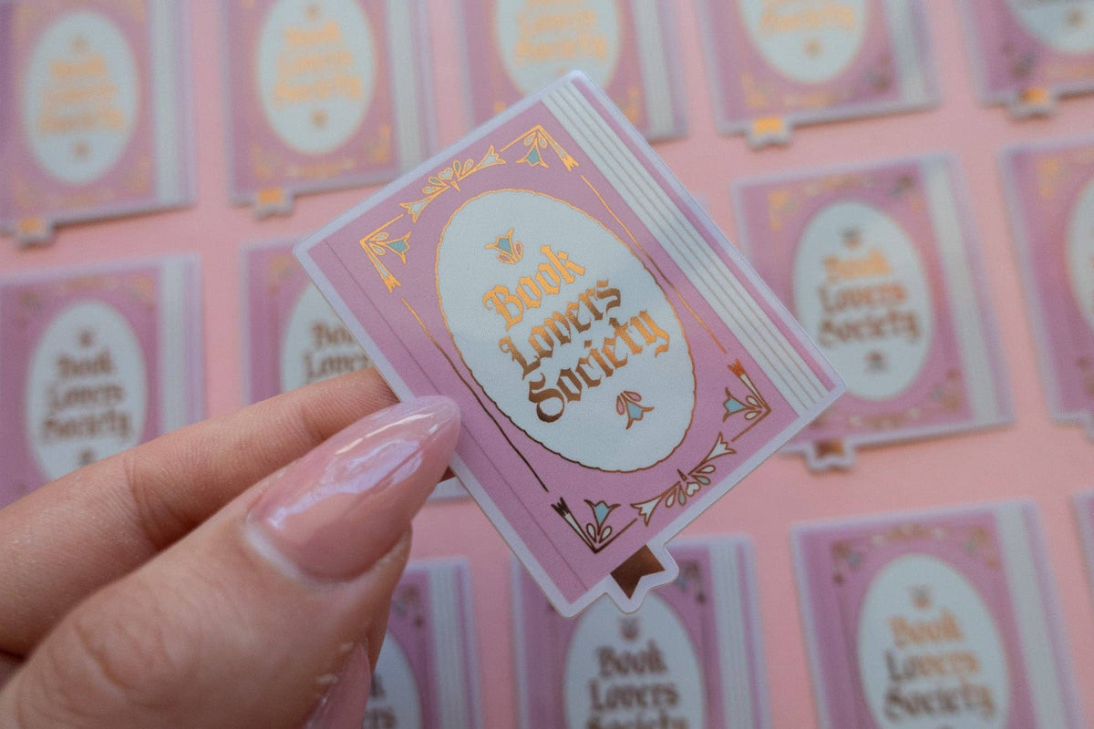 Hand holding vinyl sticker with pink frame and gold "Book Lover's Society" lettering