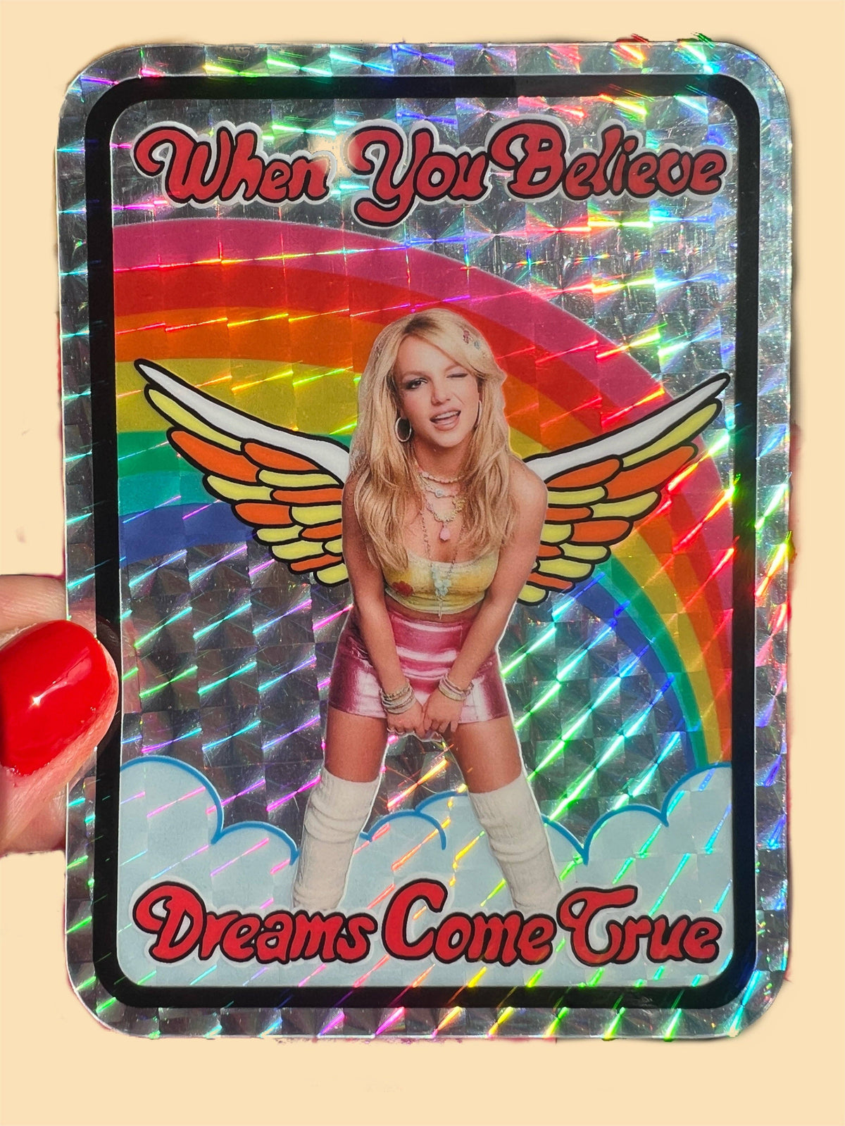 4-inch holographic sticker held by red-nailed finger, wings, and “When You Believe / Dreams Come True”.