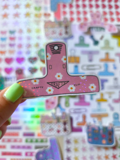 Hand holding pink floral clip sticker with "Crafty Clip" banner and flower icons