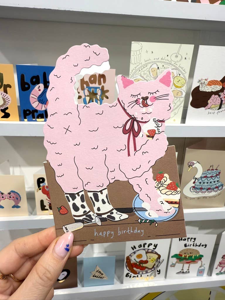 Die-cut birthday card held in hand, pink cat cake with “happy birthday”.