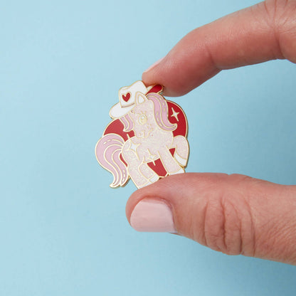 Cowgirl pony enamel pin held in fingers: white unicorn with red cowboy hat and hearts