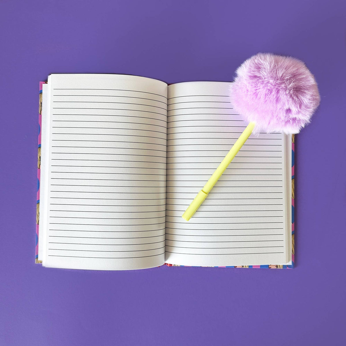 A5 notebook open with lined pages, purple cover, yellow pen with pink-purple pom.