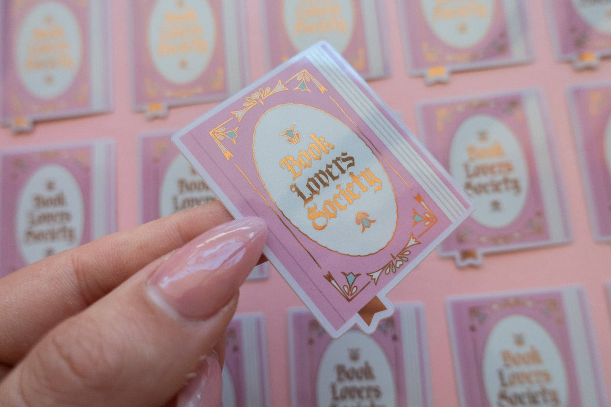 Hand holding vinyl sticker with pink frame and gold "Book Lover's Society" lettering