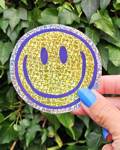 Hand holding Smiley vinyl sticker, yellow face with blue outline and glitter rainbow border.