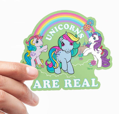 Vinyl sticker held by fingers; three unicorns under a rainbow, 'Unicorns Are Real'