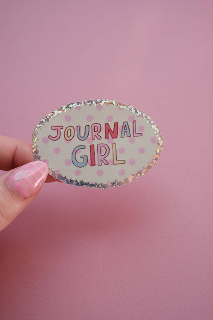 Single oval sticker reading “JOURNAL GIRL” with speckled border, held by a hand.