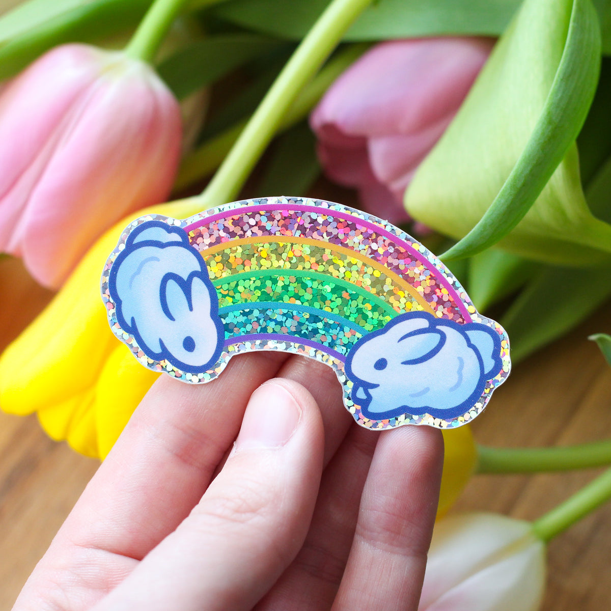 Hand holding rainbow bunny glitter sticker with arched rainbow and blue bunny head accents