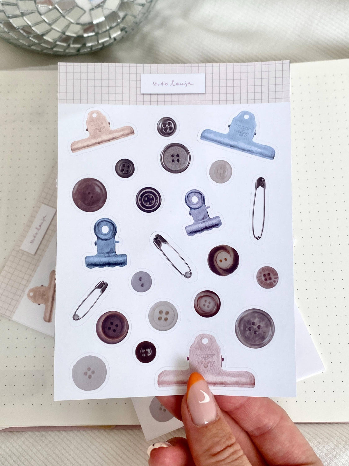 Handheld button-themed sticker sheet with blue and pink clip shapes and round buttons