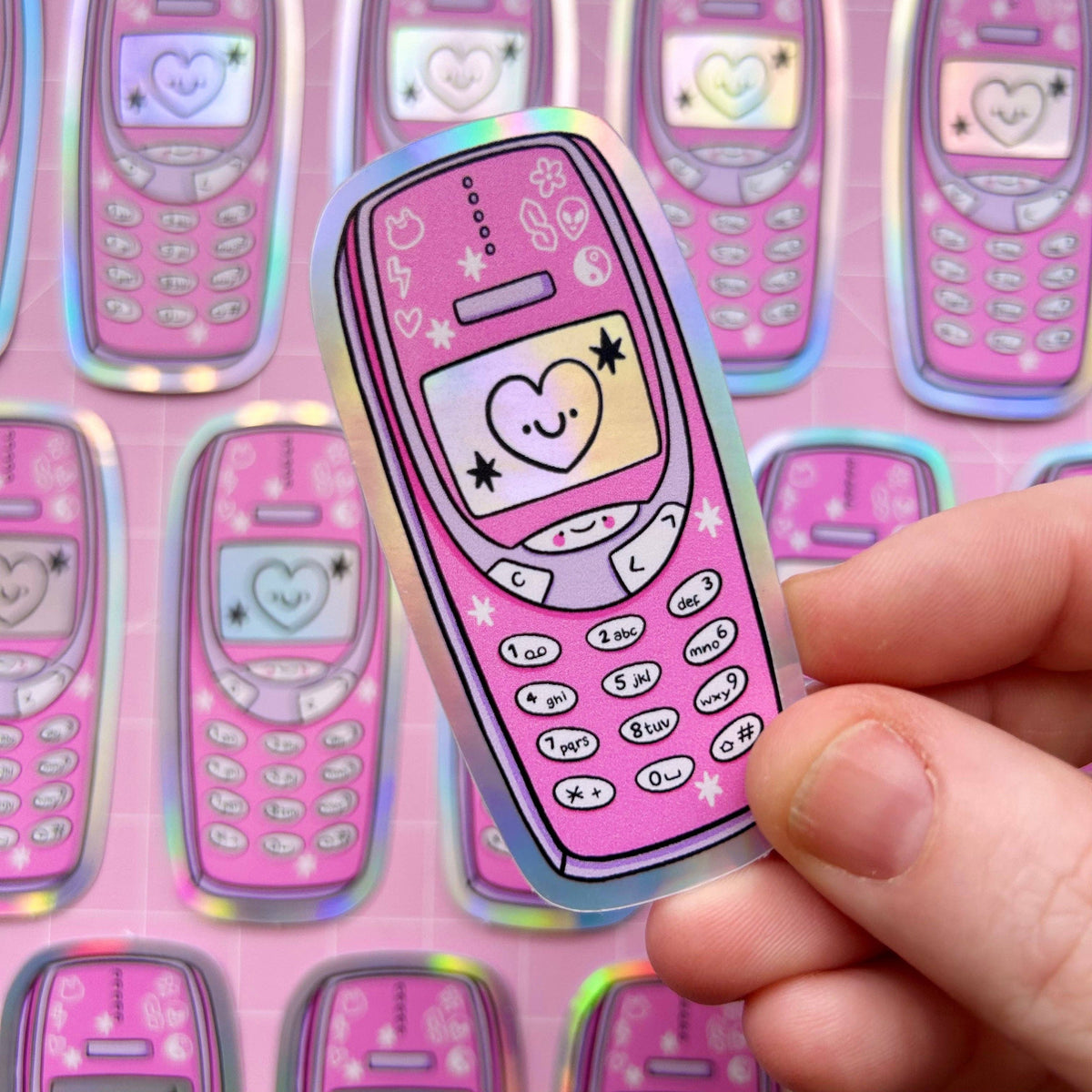 Pink holographic phone-shaped sticker with heart, smiley, stars, and button icons