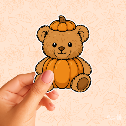 Hand holding a brown teddy bear sticker with an orange pumpkin