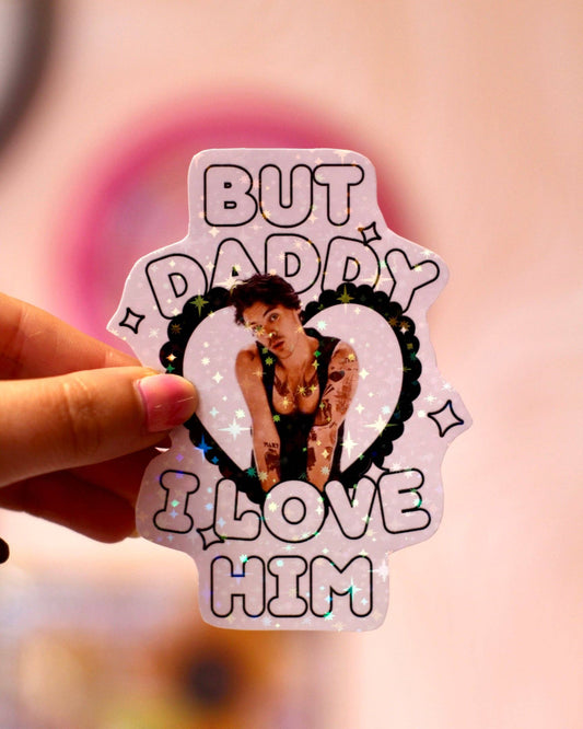 Hand holding holographic heart sticker reading 'BUT DADDY I LOVE HIM' with Harry Styles photo