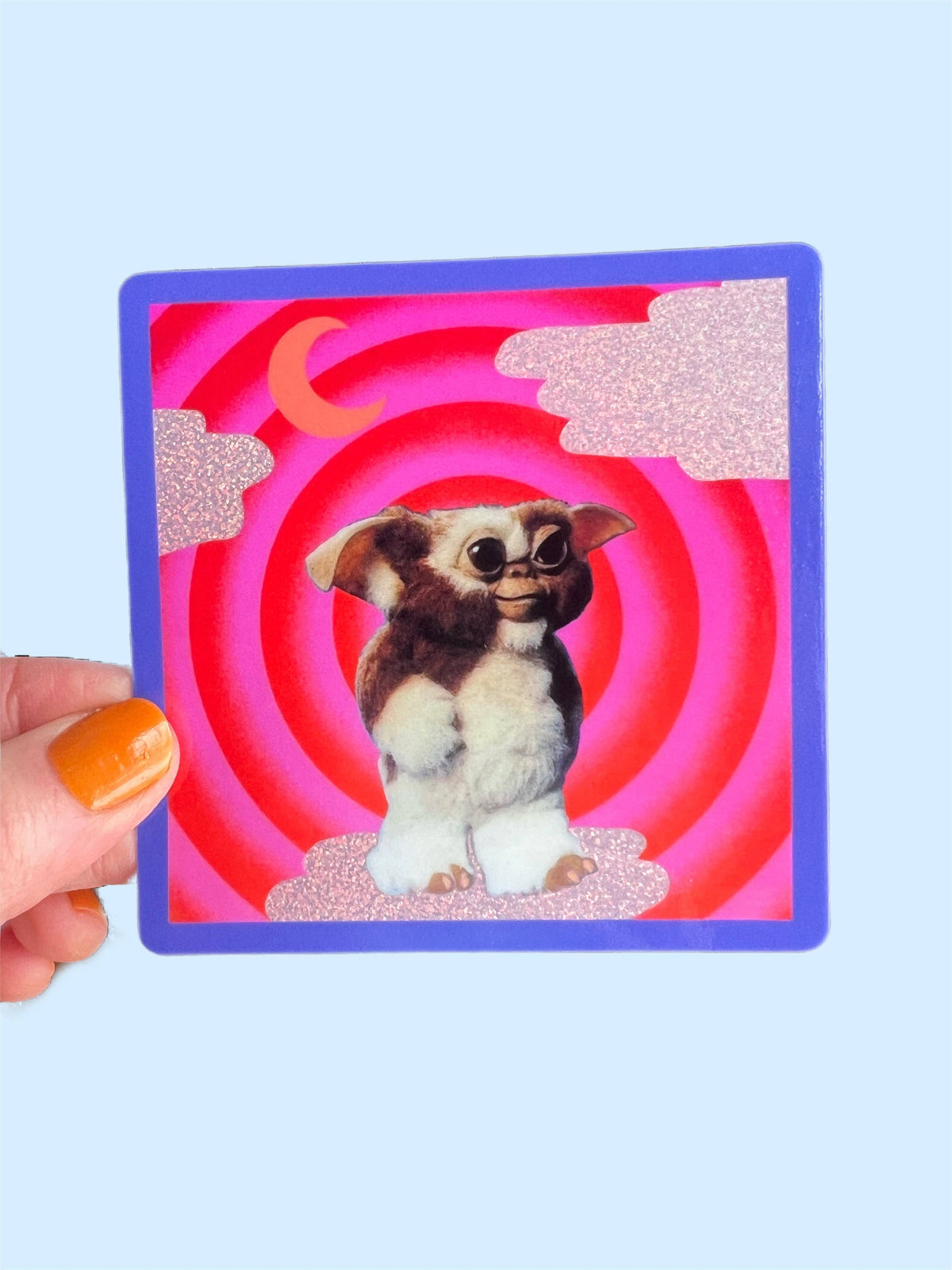 Hand holding 3-inch square Mogwai sticker, blue border and red spiral cartoon dog