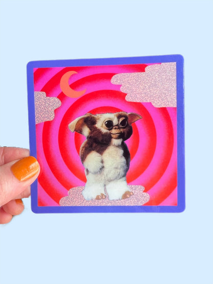 Hand holding 3-inch square Mogwai sticker, blue border and red spiral cartoon dog