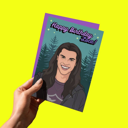 Hand holding teal birthday card with smiling vampire portrait, pine trees, and 'Happy Birthday Loca!'