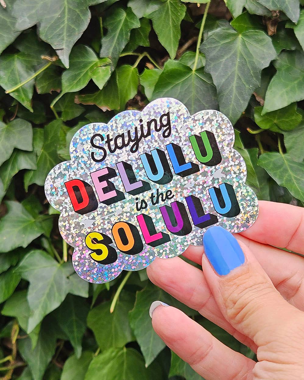 Glitter vinyl flower sticker with rainbow 'Staying Delulu' text in hand