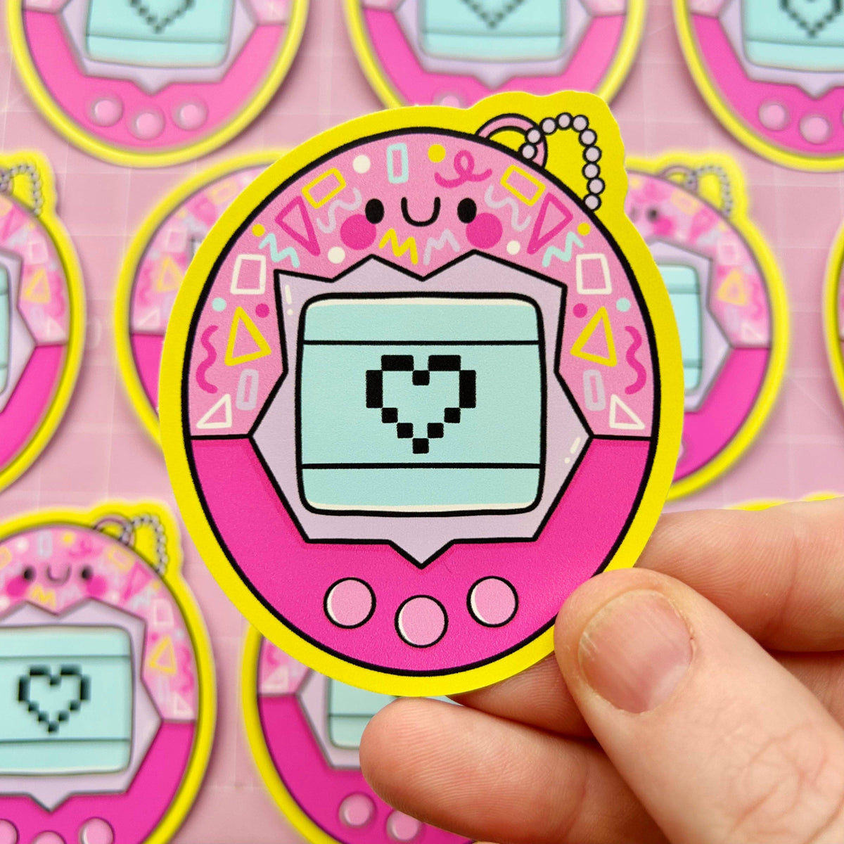 Single oval virtual pet sticker, pink border, teal display with pixel heart; held in fingers.