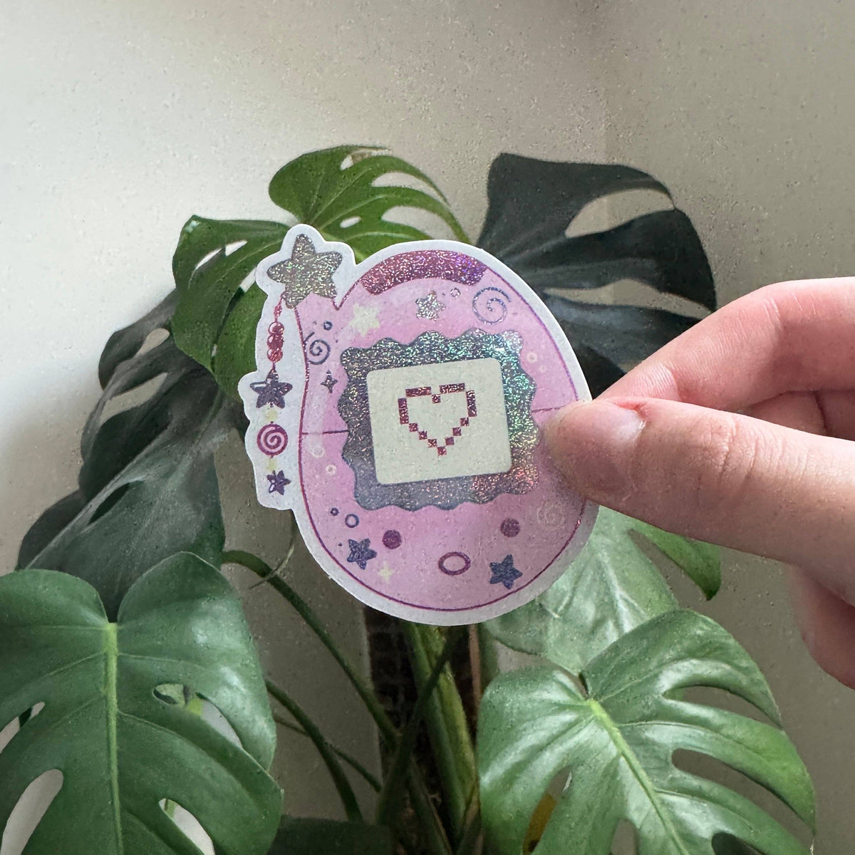 Pink oval Tamagotchi sticker with purple pixel heart, swirls, and star accents