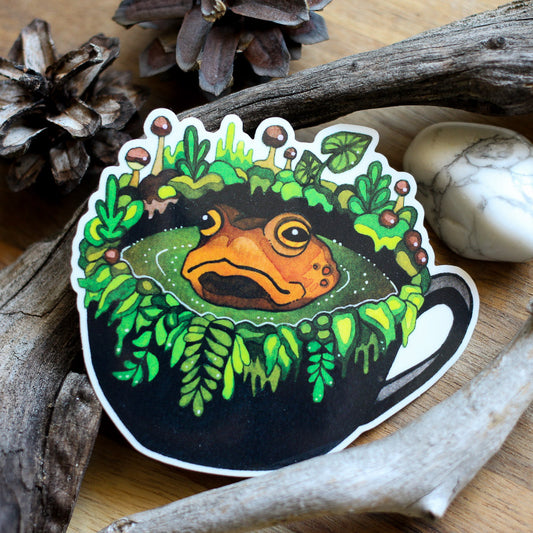 Vinyl sticker with frog face in a mug, surrounded by green leaves, white outline.